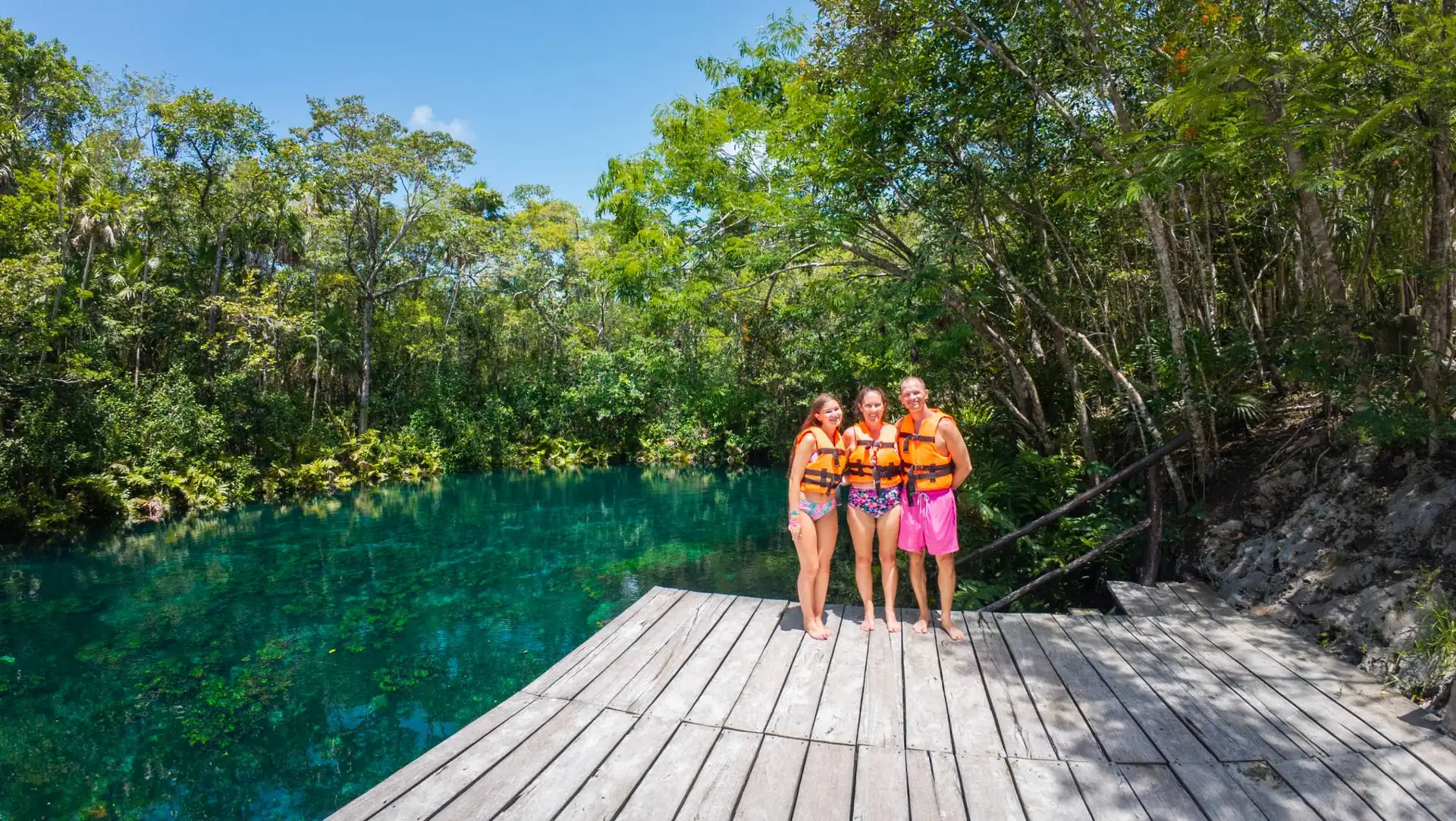 Family in life jackets on a wooden dock beside a turquoise cenote